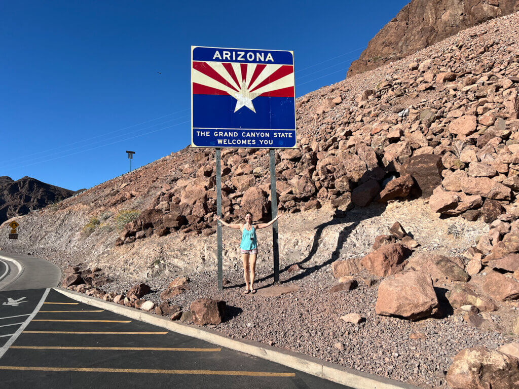 SightseeingSally stands in front of Arizona's Welcome to the Grand Canyon State sign.