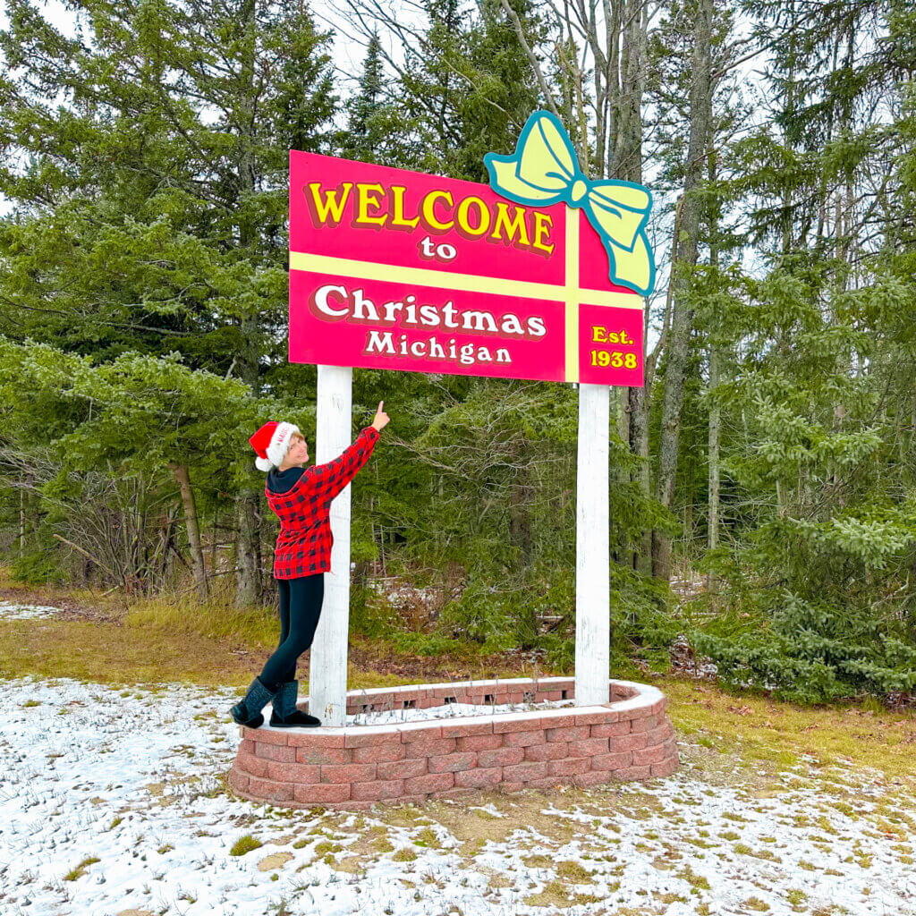 SightseeingSally wearing a Santa cap stands by the welcome to Christmas Michigan sign.