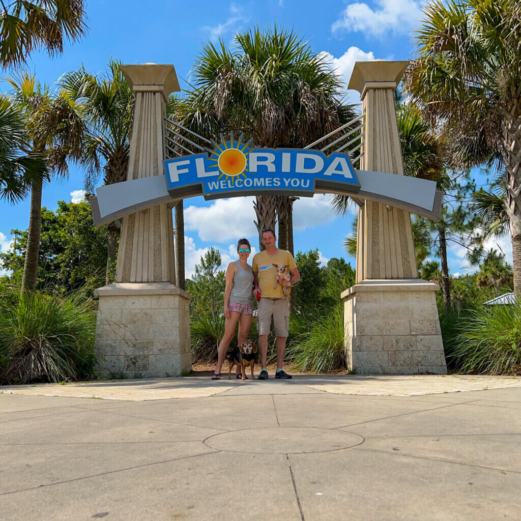 SightseeingSally along with MeanderingMarty and their two dogs are standing beneath a Florida Welcomes You sign at a rest stop on the Florida border.