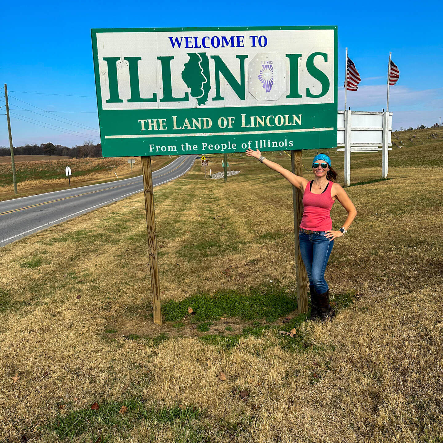 SightseeingSally stands in front of a Welcome to Illinois sign beside a two lane country road.