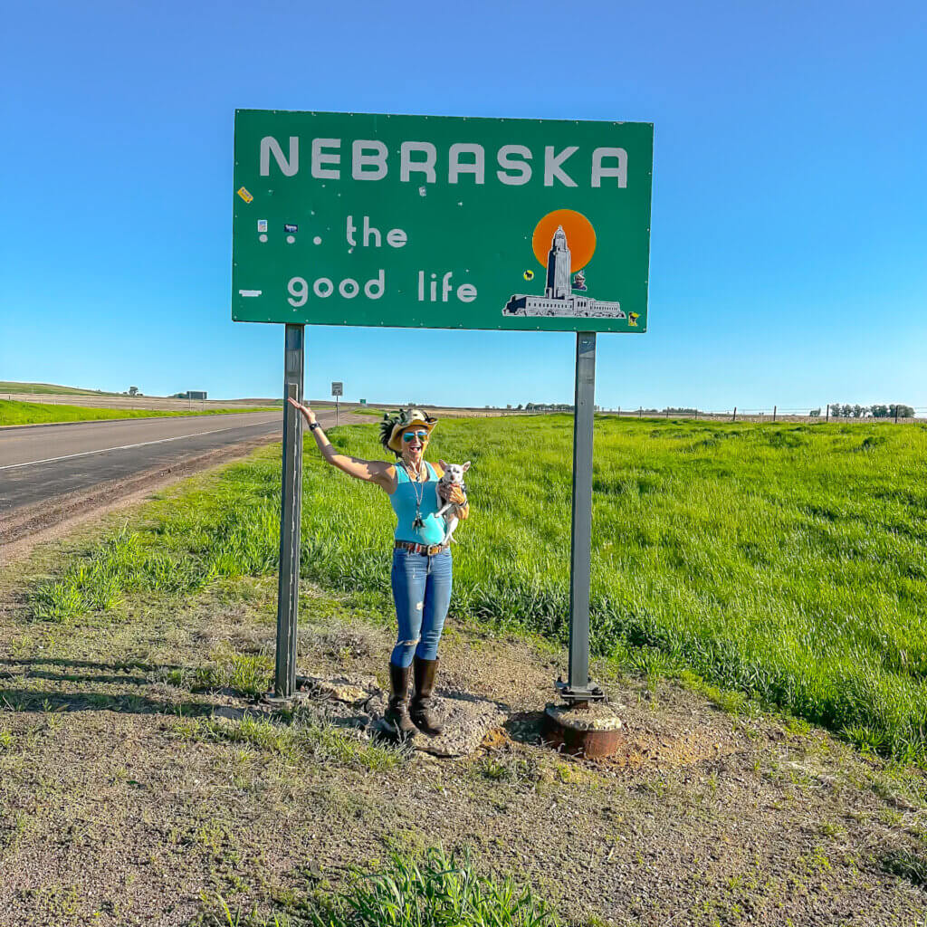 SightseeingSally stands in front of Nebraska's state sign  along side a deserted country highway while holding her dog Leo, a white chihuahua.