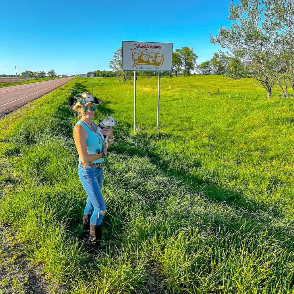 SightseeingSally holding a white chihuahua standing in front of a South Dakota welcome sign beside a country road.