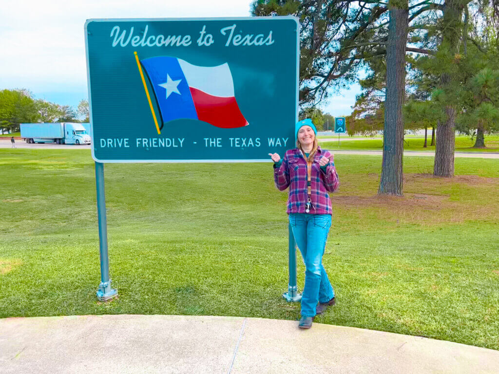 SightseeingSally stands next to a Welcome to Texas sign at a welcome center in Texas.